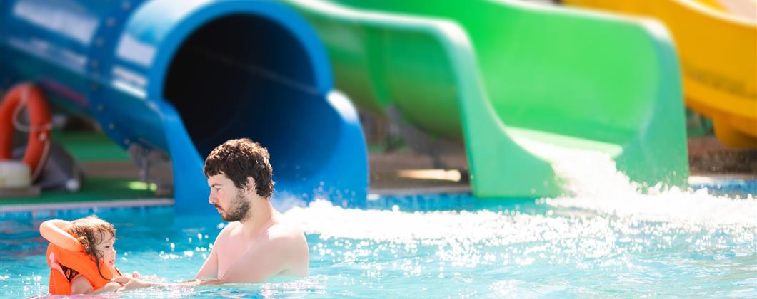 A parent plays with their child who is wearing an inflatable jacket in a water park pool, colourful slides are in the background